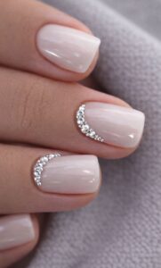 Short nude glossy nails with curved rhinestone accents near the cuticle on a soft neutral background.