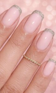 Short natural nails with soft pink polish and silver glitter French tips on a hand with a delicate gold ring.