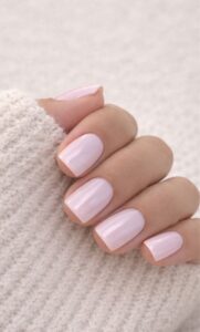 Soft pink short square nails with a glossy finish, styled with a cream knit sweater and neutral blurred background.