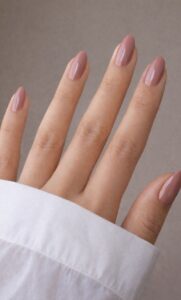 Dusty pink almond-shaped nails on a hand with a white shirt sleeve and soft neutral background.